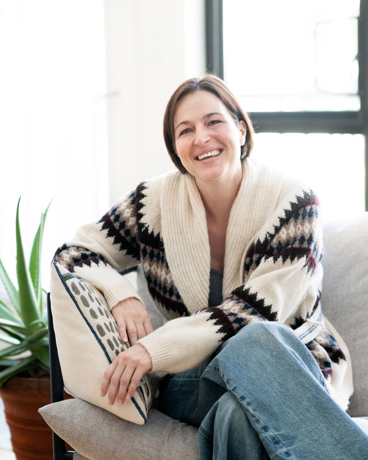 Pamela Wallberg laughing on a sofa beside a plant, wearing a patterned cardigan